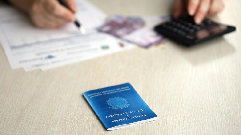 Carteira de Trabalho azul sobre mesa clara, ao lado de documentos, cédulas de dinheiro e mãos usando calculadora.
