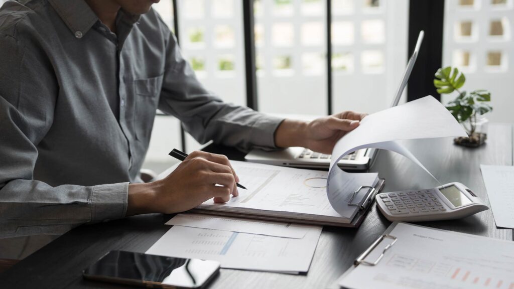 Pessoa analisando relatórios financeiros, com calculadora, notebook e planilhas sobre a mesa, em atividade relacionada a empreender no mercado financeiro.