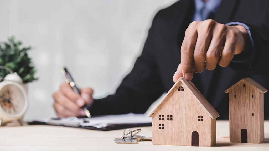 Pessoa em traje formal segurando uma miniatura de casa sobre uma mesa, ao lado de chaves e documentos, representando a contratação de um empréstimo com garantia de imóvel.