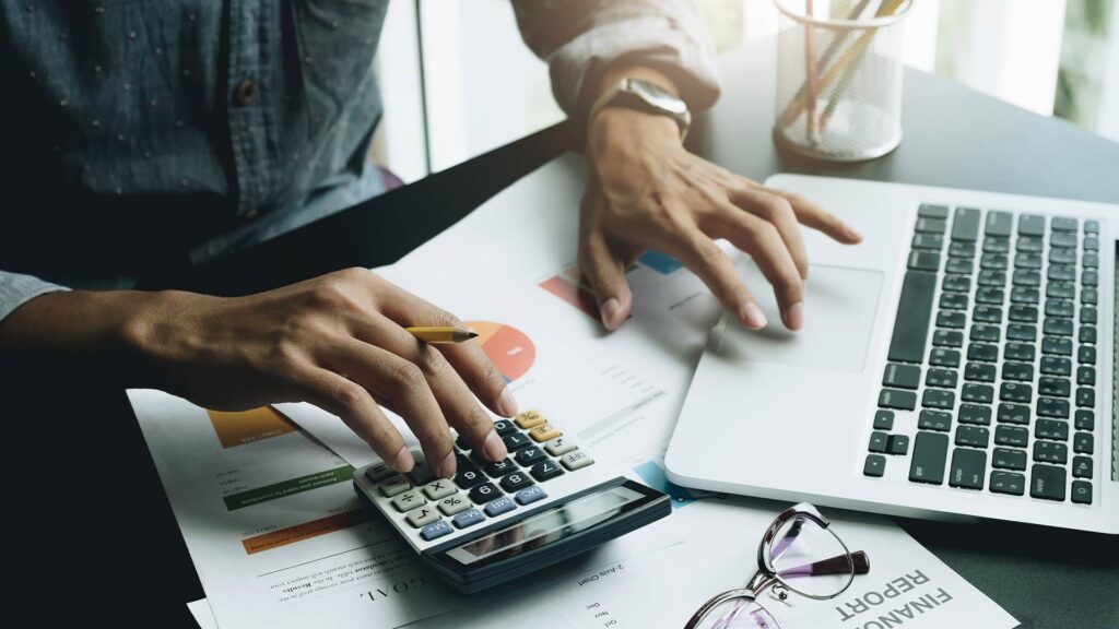 Pessoa utilizando calculadora e notebook ao revisar relatórios financeiros e documentos, em atividade relacionada à declaração do Imposto de Renda.