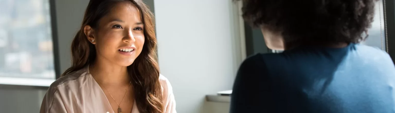 Duas pessoas em uma conversa profissional em um ambiente iluminado por luz natural. A mulher à esquerda, com cabelos longos e castanhos, sorri enquanto fala, transmitindo simpatia e engajamento na interação