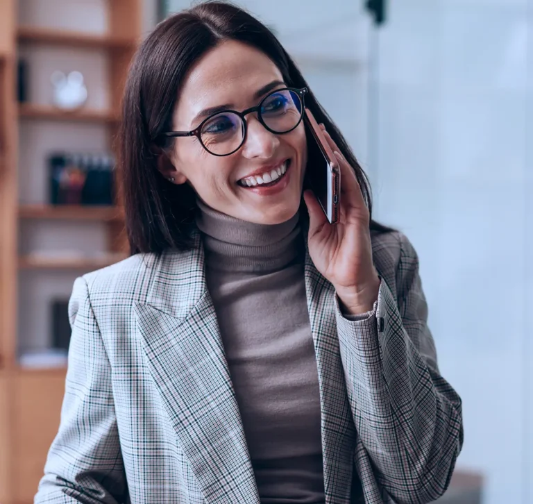 Mulher sorridente de óculos e blazer cinza falando ao celular em um ambiente de escritório.