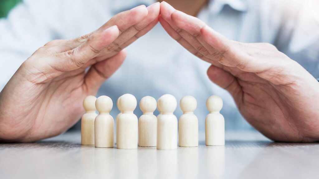 Mãos em formato de proteção cobrindo figuras humanas de madeira, simbolizando a proteção oferecida por um seguro de responsabilidade civil empresarial.