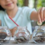 Criança organiza moedas em três potes de vidro sobre uma mesa, representando a divisão do Planejamento Financeiro.