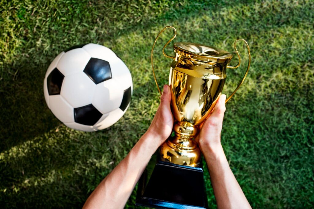 Bola de futebol ao lado de uma taça sendo segurada por um par de mãos, representando Seguro para atletas dentro e fora de campo.