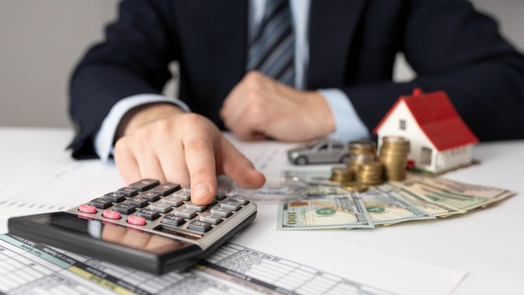 Homem de terno usa calculadora sobre mesa com miniatura de casa, carro, moedas e notas. Imagem representa o planejamento de empréstimo e financiamento.