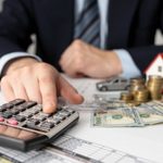 Homem de terno usa calculadora sobre mesa com miniatura de casa, carro, moedas e notas. Imagem representa o planejamento de empréstimo e financiamento.